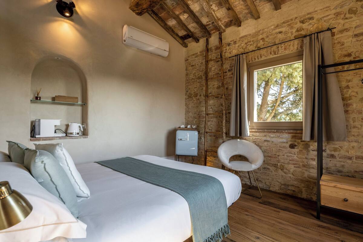 A bedroom with a bed, a bedside table, a chair, a window, and a stone wall.
