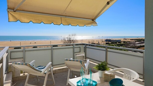 A balcony with a view of the beach, white chairs, a table with a potted plant, and a beige canopy. - Beachside house with panoramic view (Bibione)