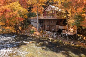 A cabin with a wooden deck overlooking a river, surrounded by autumn foliage. - Cozy 2BR Sevierville Cabin Retreat with Hot Tub, Fire Pit, and Riverfront Access (Sevierville)
