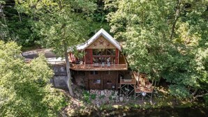 Una cabaña de madera con balcón, ubicada junto a un río rodeado de un bosque denso.