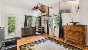 Una habitación amplia con una escalera de madera, un gran ventanal con cortinas, un aparador de madera y un televisor de pantalla plana en la pared.