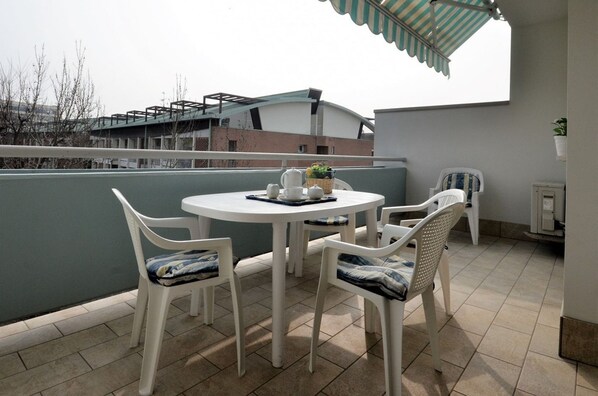 A small outdoor patio with a round table and four chairs, a small table with a teapot and cups, and a striped awning. - Lovely apartment with pool and solarium (Bibione)