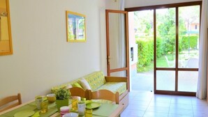 A cozy room with a dining table set for two, a sofa, and a sliding glass door leading to a garden. - A green escape in Bibione (Bibione)
