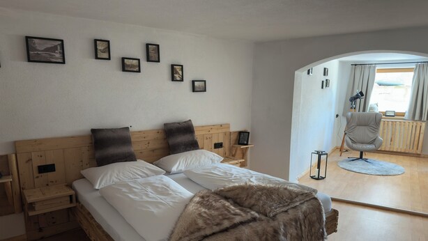 Camera da letto con una testiera in legno, biancheria bianca, una coperta in pelliccia e vista su un balcone con una sedia e un tavolo.