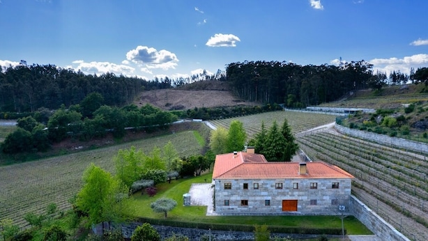 A stone building with a red roof surrounded by vineyard rows, set in a hilly landscape.