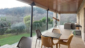 A covered outdoor patio with a table and chairs, a telescope, and a view of a hilly landscape.