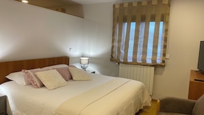 A hotel room with a large bed, a nightstand with a lamp, a chair, a window with striped curtains, and a television on a wooden stand.