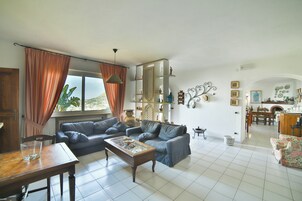 Amplia sala de estar con sofá, mesa de centro y mesa de comedor. Se ve un área de cocina al fondo.