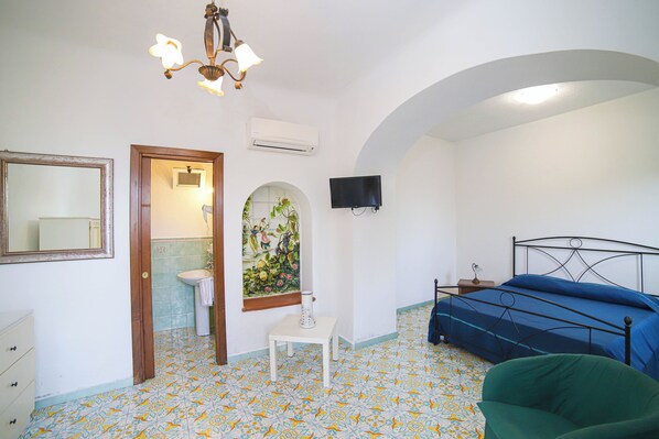 A bedroom with a bed, a chair, a TV, a painting, a sink, and a mirror.
