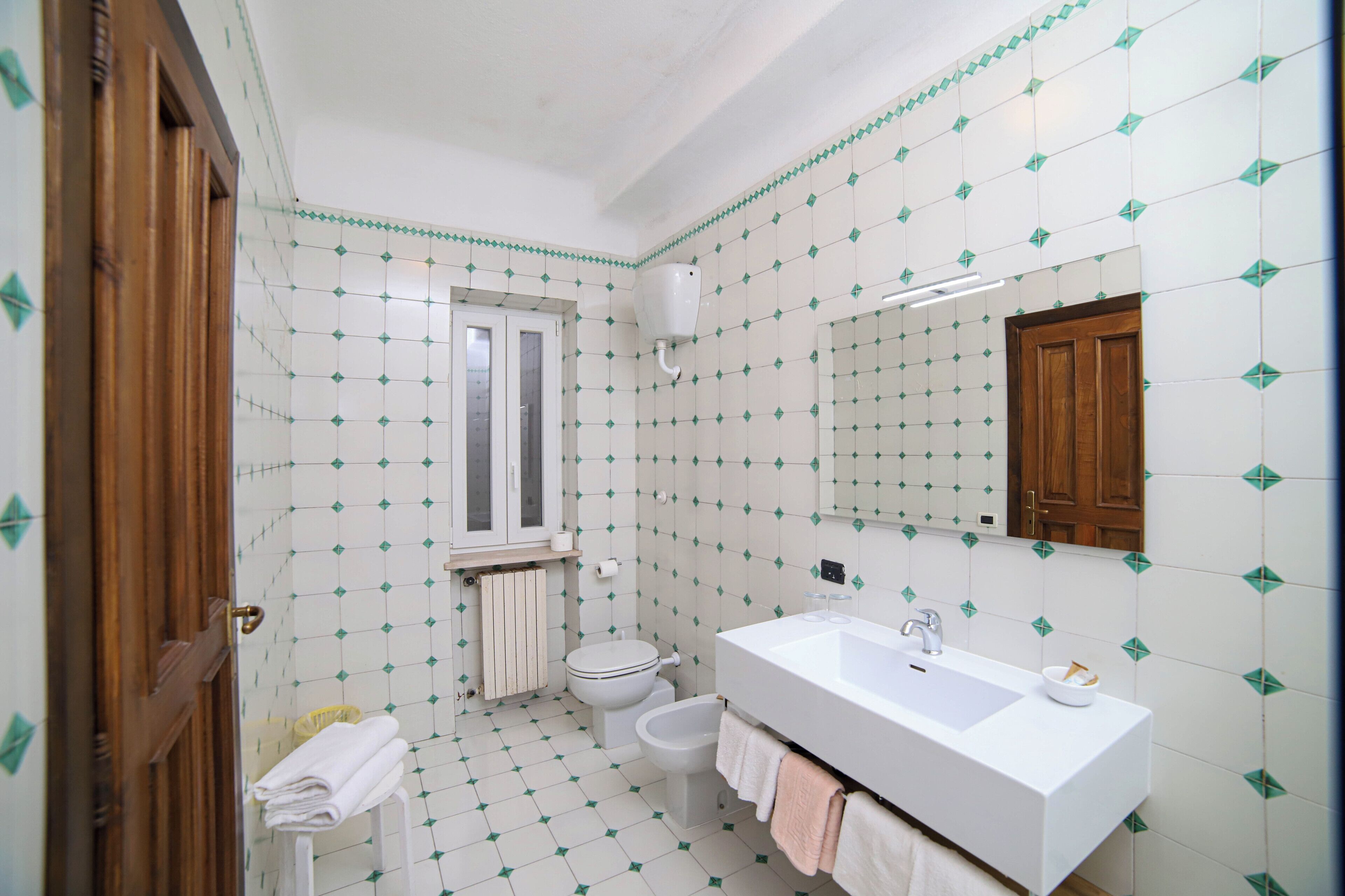A bathroom with a white sink, toilet, and bidet, tiled floor, and a wooden door.