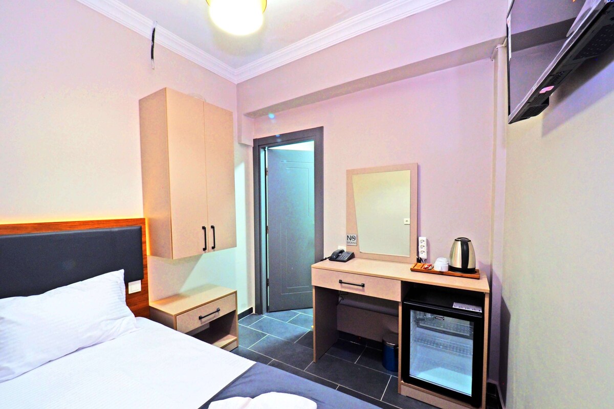A hotel room with a bed, a desk with a computer, a minibar, and a door.