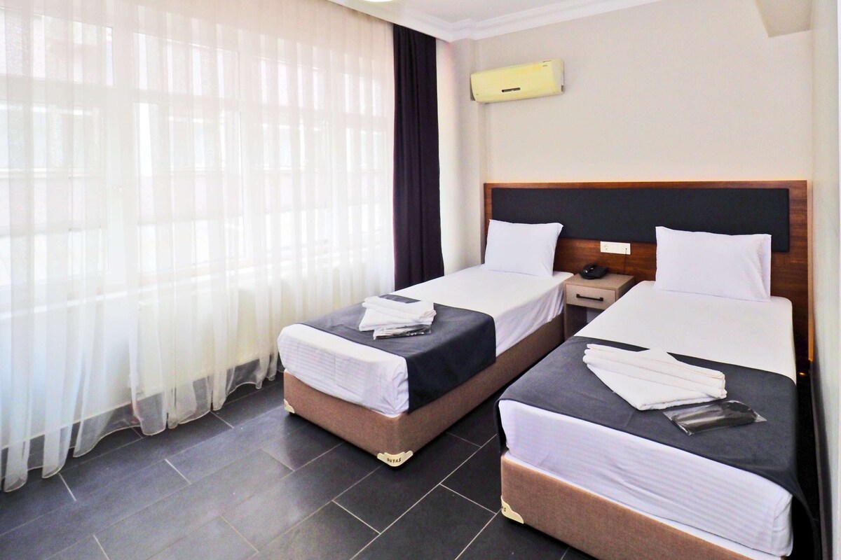 A hotel room with two beds, a bedside table with a phone, and a window with sheer curtains.