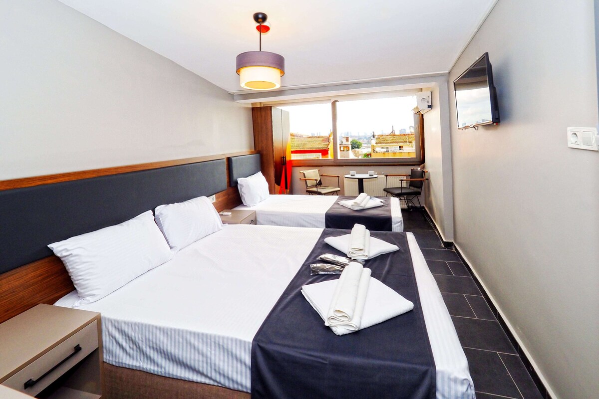 A hotel room with two beds, a desk, a chair, a small table, and a window with a city view.