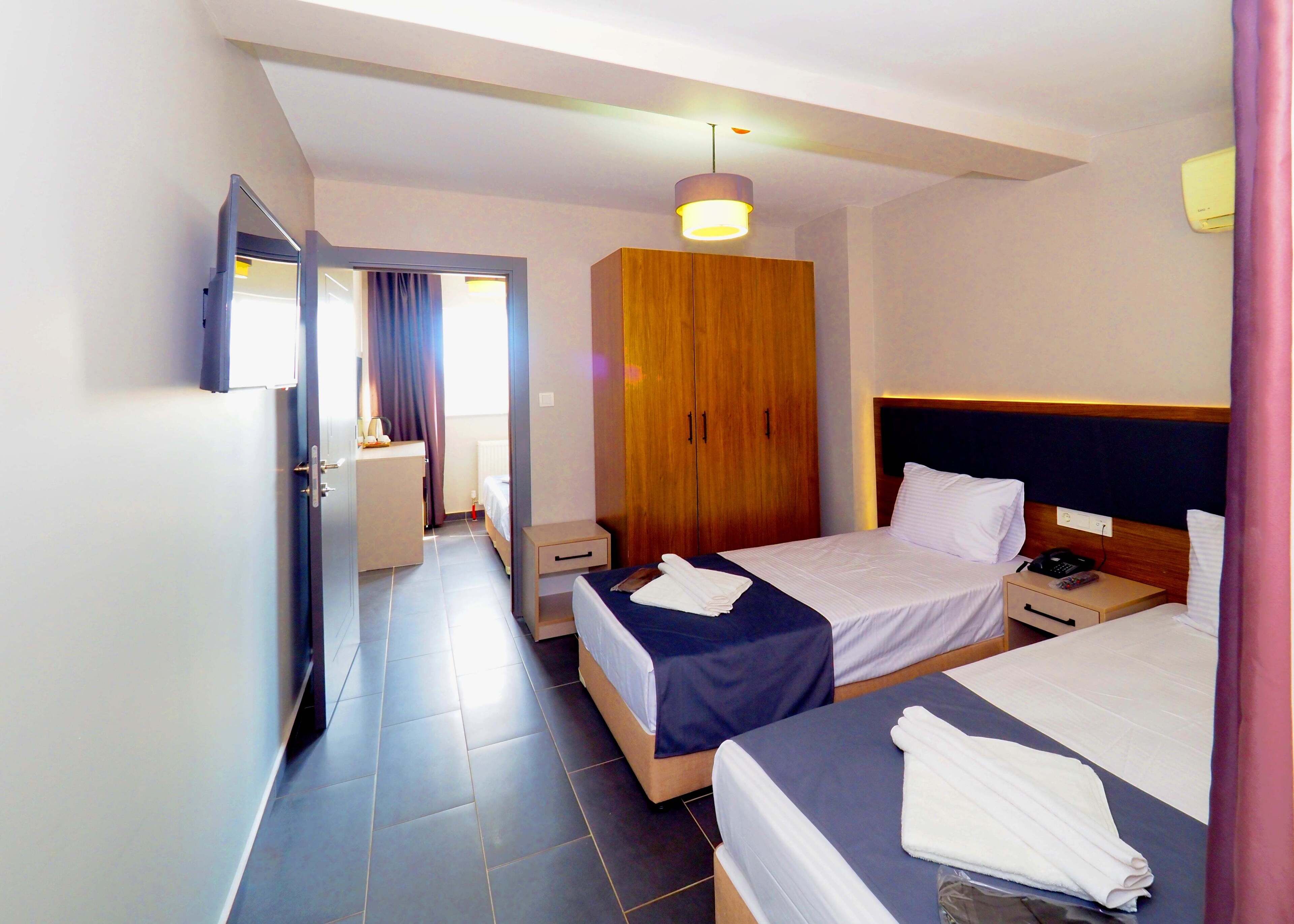 Quarto de hotel com duas camas, um guarda-roupa de madeira, uma secretária com abajur e um banheiro visível através de uma porta aberta.