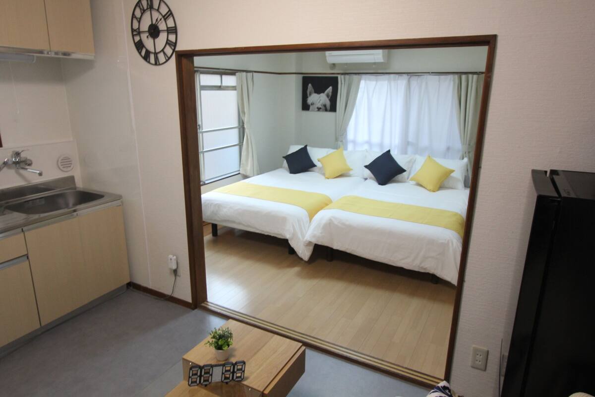 A compact hotel room with a mirrored wardrobe, a bed with two pillows, a small table with a plant, and a visible kitchen area.