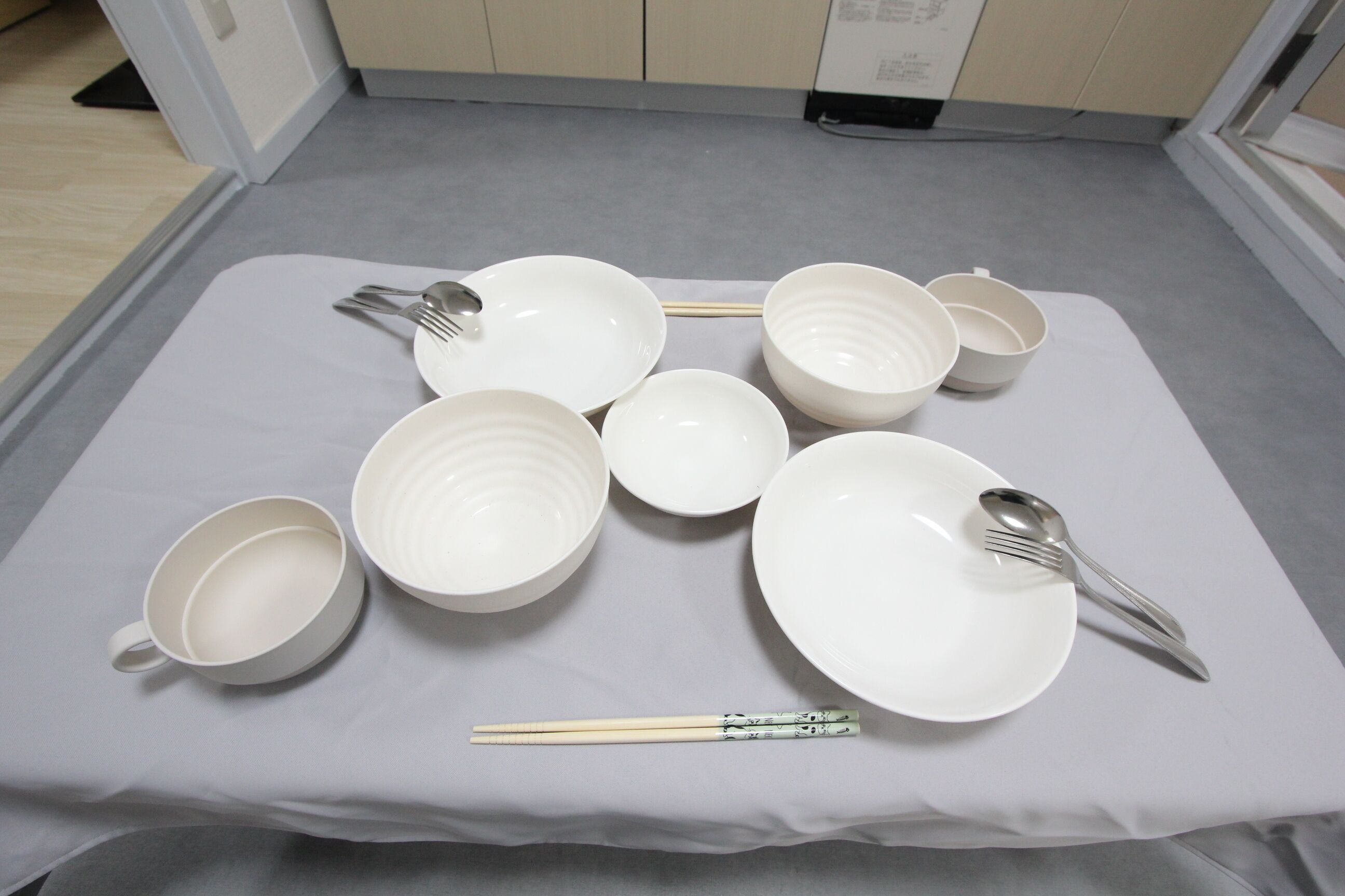 A neatly arranged table with white plates, bowls, and chopsticks on a white tablecloth in a room with light-colored walls and a gray carpet.