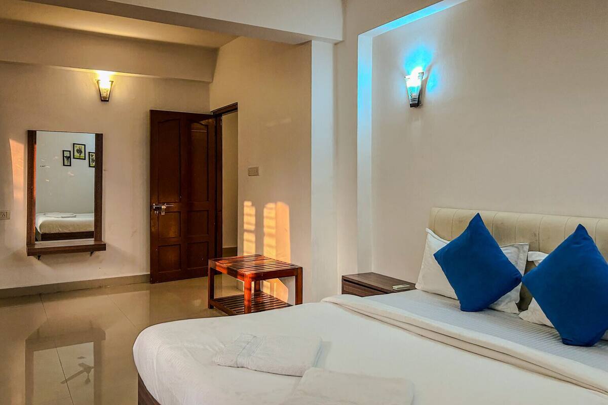 Habitación de hotel con una cama, una mesita de noche, una silla de madera, un espejo y una lámpara fijada a la pared.