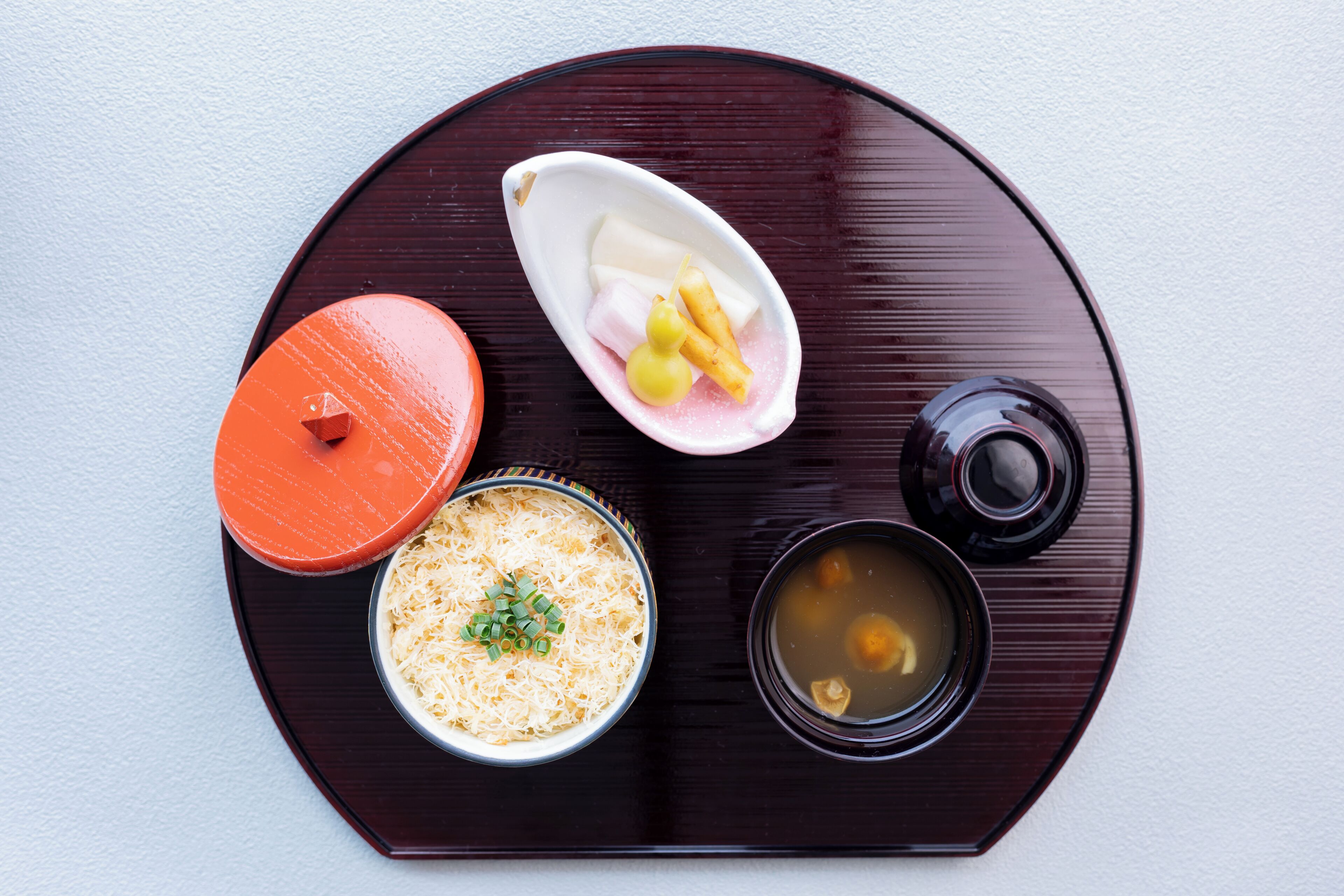Una bandeja redonda con un tazón de fideos cubiertos de cebolletas, un recipiente pequeño con vegetales encurtidos, un tazón de sopa y un plato pequeño con un trozo de carne.