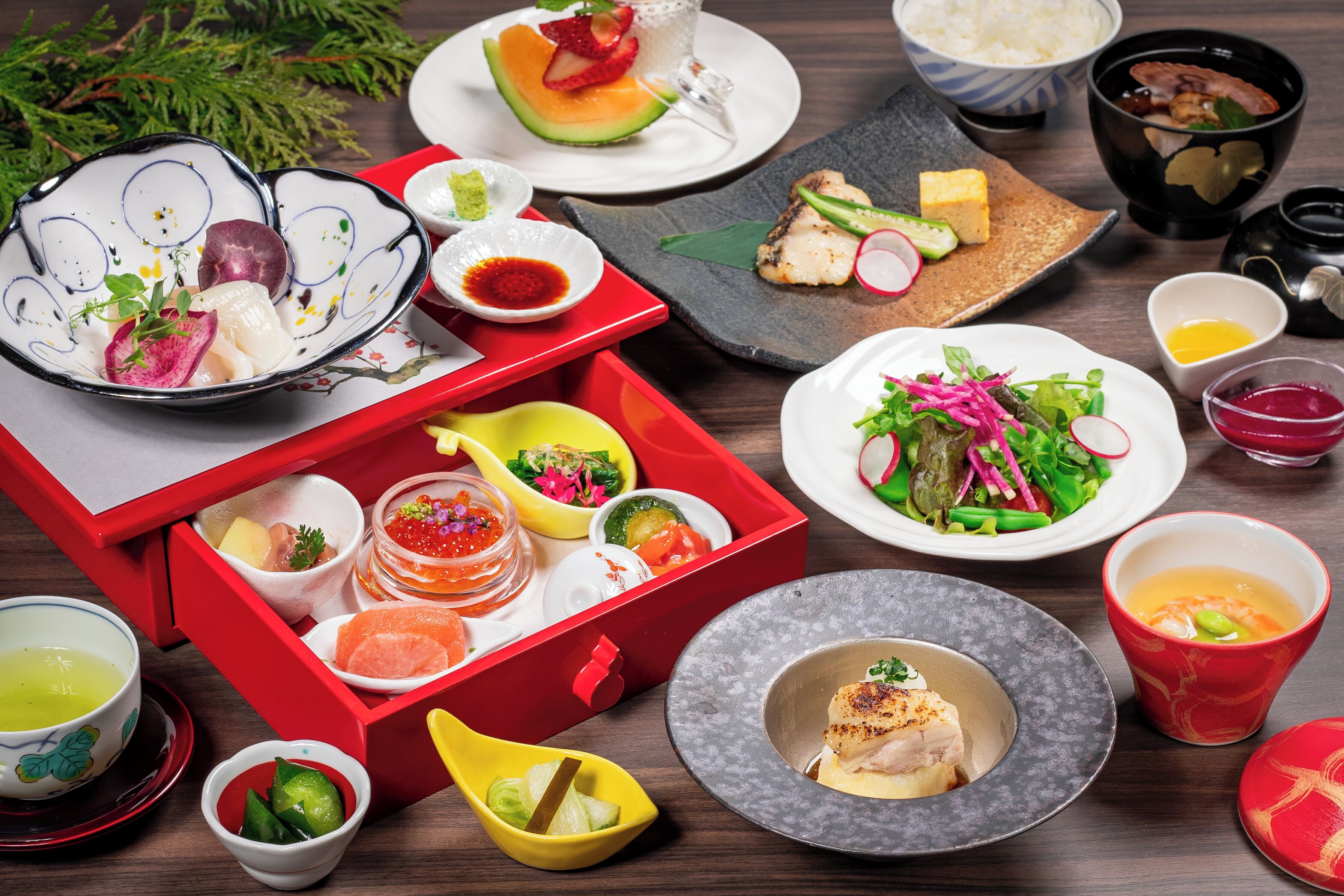Variedad de platos y alimentos dispuestos sobre una mesa, incluyendo una caja roja con comida, tazones, platos y una taza.