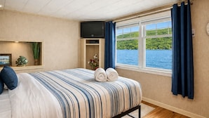 Ein Hotelzimmer mit Bett, Fernseher, Fenster mit Vorhängen, einer Pflanze und Blick auf einen See.