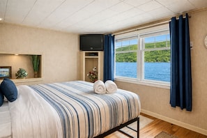 A hotel room with a bed, a TV, a window with curtains, a plant, and a view of a lake.