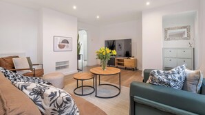 A modern living room with a sofa, a coffee table, and a TV. There is a dining area with a table and chairs, and a kitchen in the background. - Bluebell House - Sleeps 5 - Garden - Parking (Peterborough)