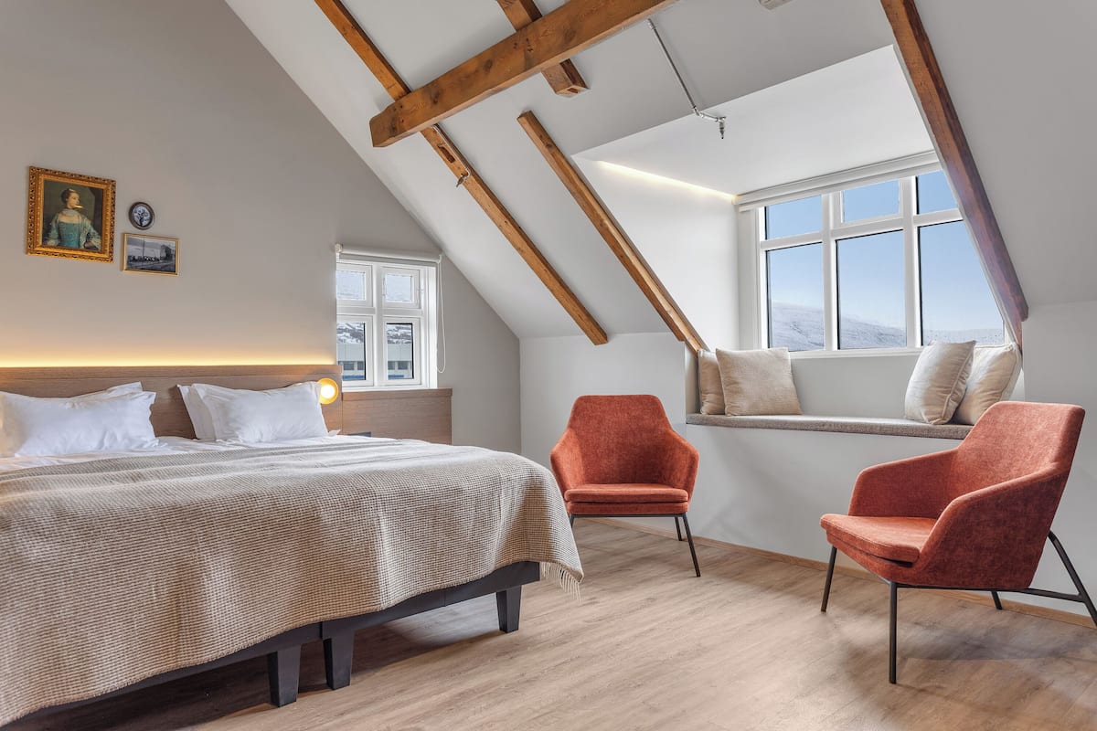 A bedroom with a sloped ceiling, a bed, a window seat, and two chairs.