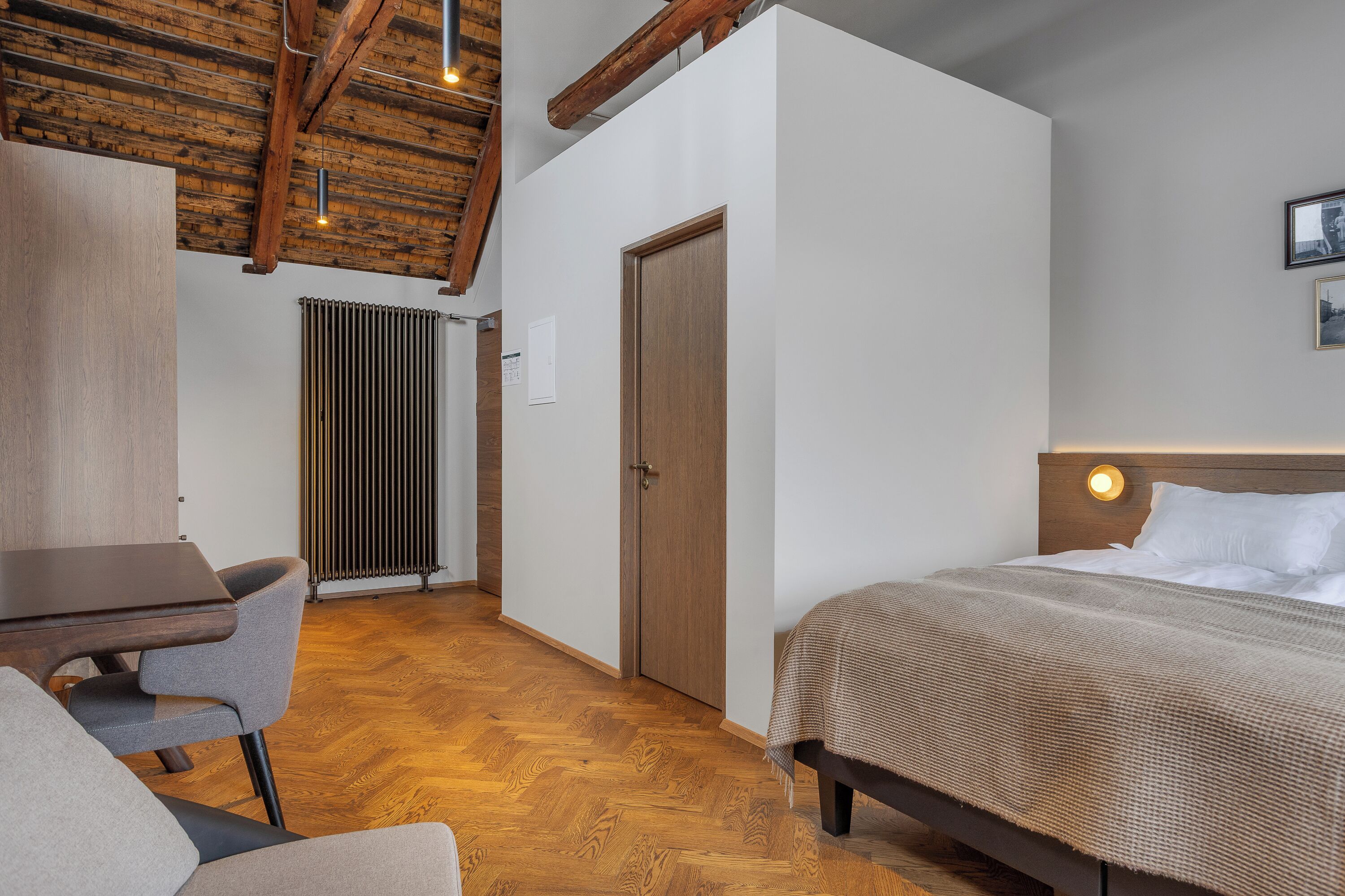 A room with a bed, a desk, and a chair. There is a radiator, a door, and a light fixture on the wall.