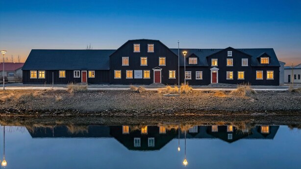 A large, dark-colored building with multiple windows and a red door, reflecting on a calm body of water. - Bryggjan Boutique Hotel (Akureyri)