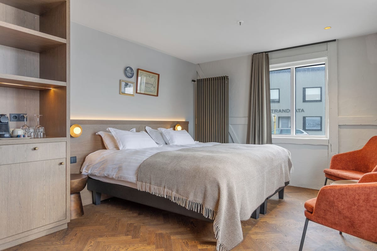 A hotel room with a bed, a wooden wardrobe, a bedside table, a chair, and a window with curtains.