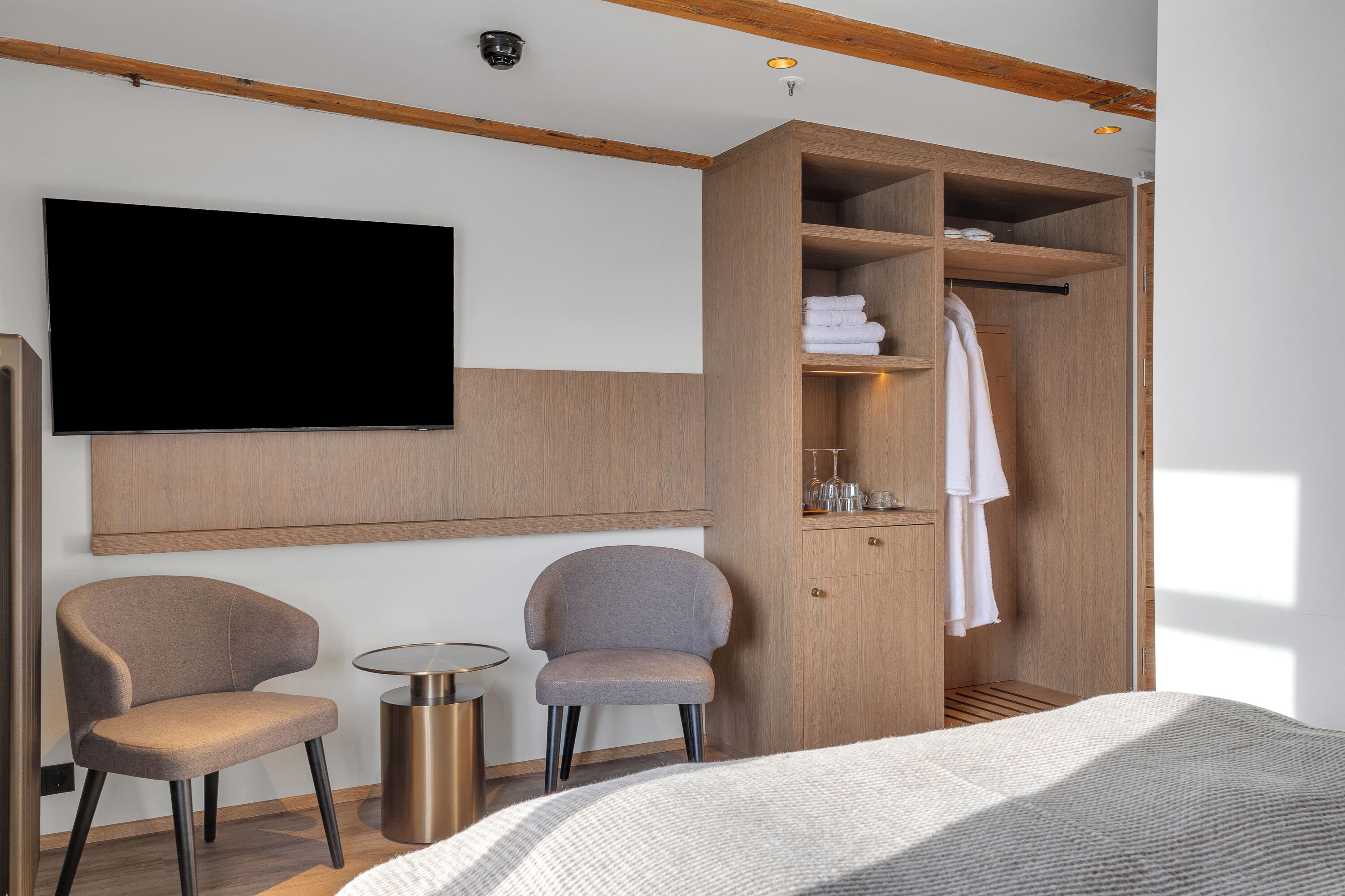 Una habitación de hotel moderna con una cama grande, dos sillones, un televisor montado en la pared, un armario de madera y una pequeña mesa redonda.
