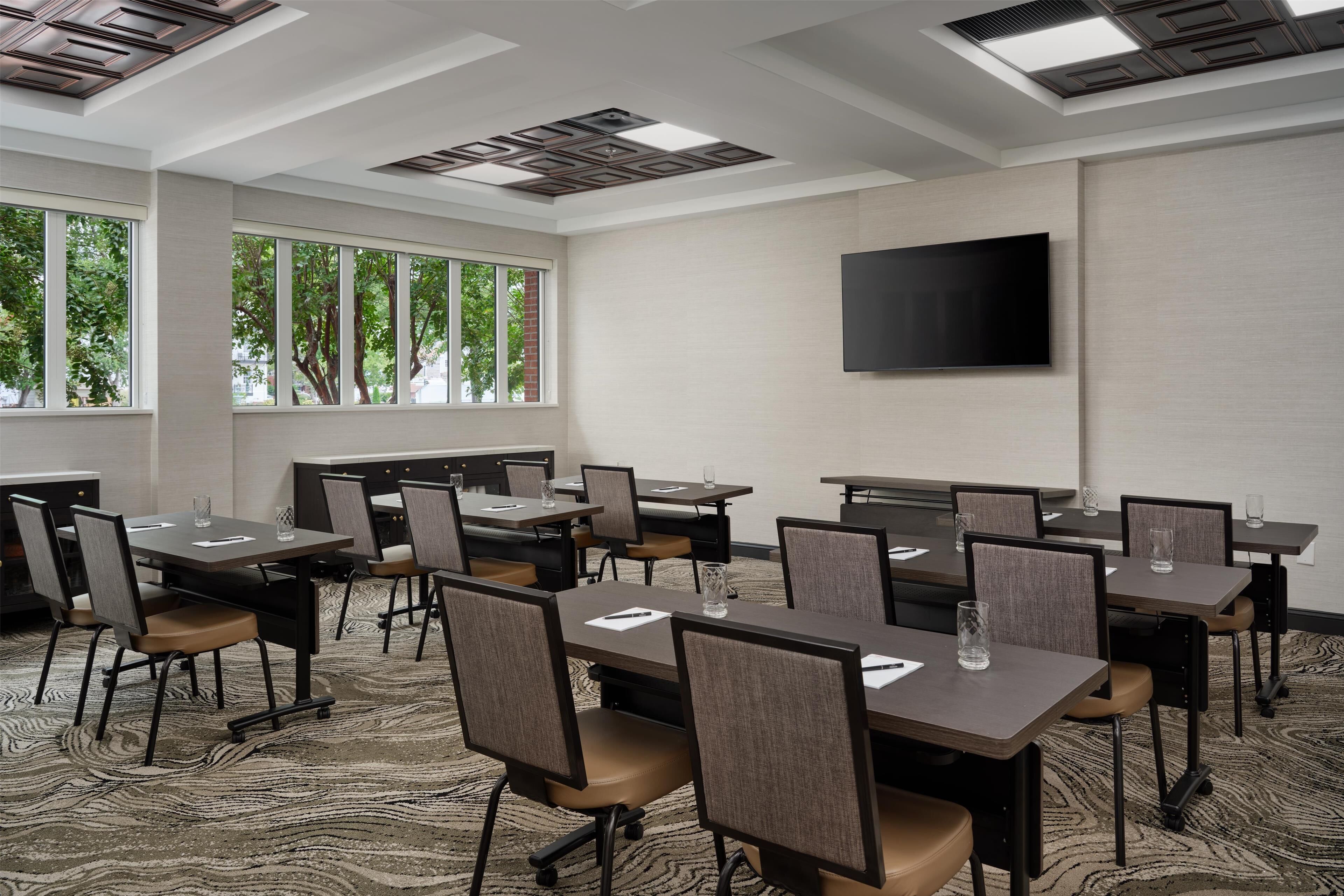 Una sala conferenze con diversi tavoli e sedie, ampie finestre con vista sugli alberi, una TV a schermo piatto montata a parete e un tappeto decorato.