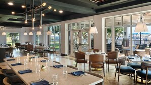 A restaurant with a long communal table, individual tables, and chairs. There are hanging pendant lights and a ceiling with a geometric pattern. - Tryon Riverfront Inn New Bern, Tapestry (New Bern)