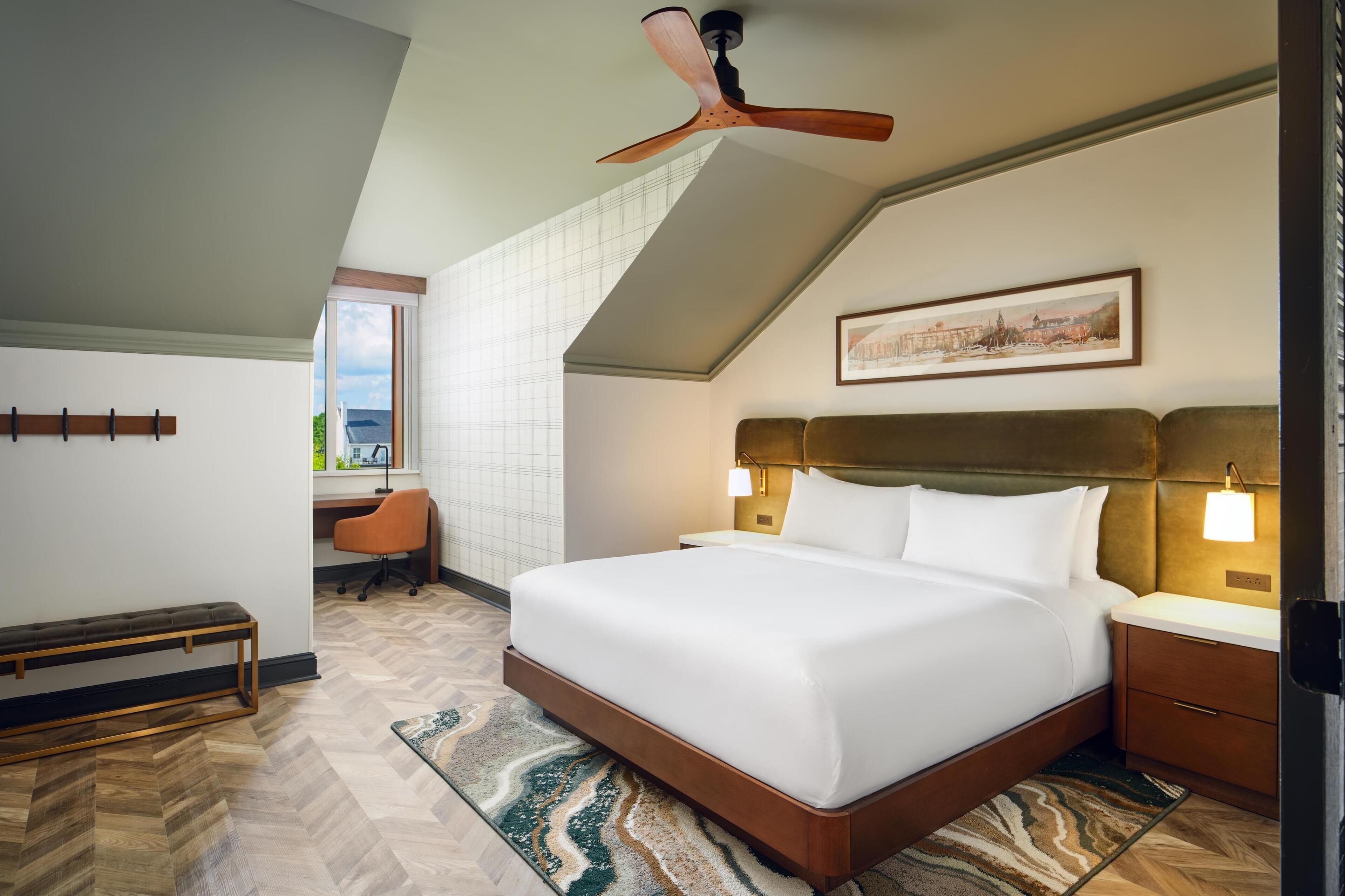 Camera d'albergo con un letto grande, una scrivania, una poltrona arancione, una panca, una ventola a soffitto e un dipinto con vista sulla città.