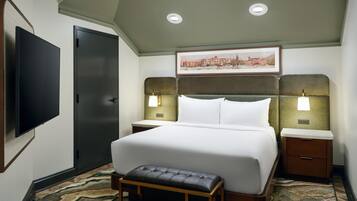 A hotel room with a large bed, a flat-screen TV, a door, two bedside lamps, a framed picture on the wall, and a bench.