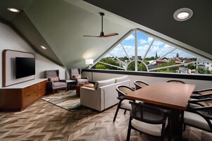 A modern loft bedroom with a large skylight, a flat-screen TV, a sofa, a dining table, and a view of a cityscape.