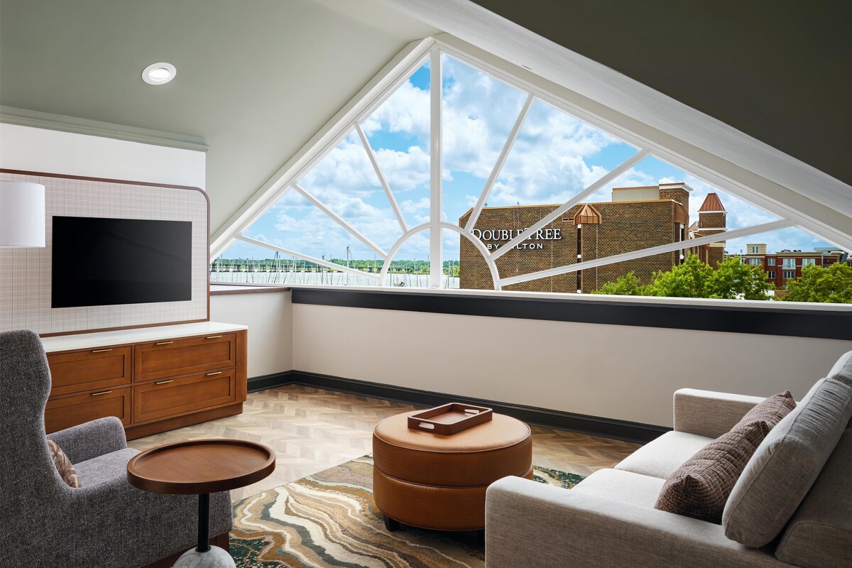 A modern living room with a large window, a flat-screen TV, a sofa, a chair, a small table, and a cabinet.