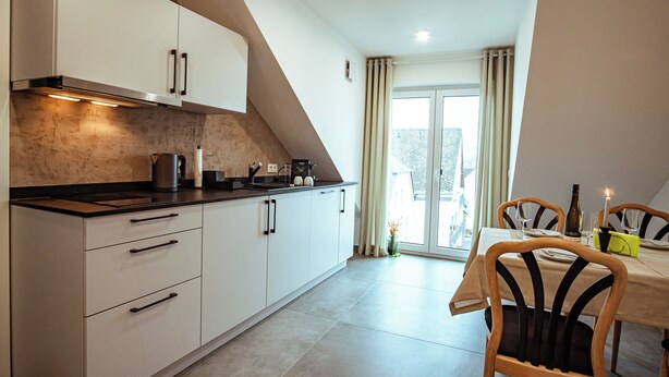 A modern kitchen with white cabinets, a black countertop, and a dining area with wooden chairs and a table. - Ferienwohnungen Moselblick Bekond (Bekond)