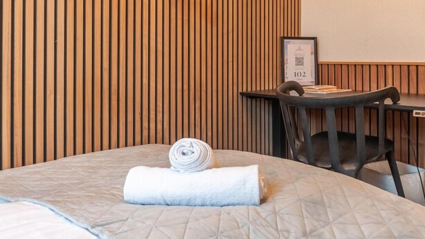 A hotel room with a bed, a desk, and a chair. There is a wooden slat wall behind the bed and a framed certificate on the desk. - Hotel Assens (Assens)