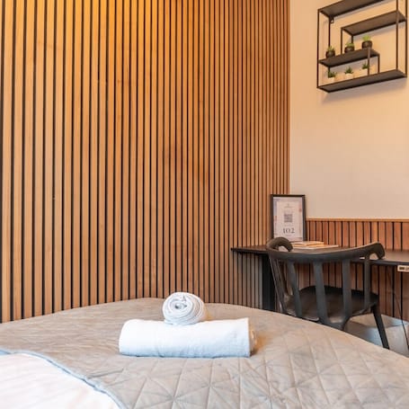 A hotel room with a bed, a desk, and a chair. There is a wooden slat wall behind the bed and a framed certificate on the desk.