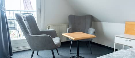 Habitación de hotel con cama, dos sillones grises, una mesita de madera, una mesita de noche blanca y ventana con vistas al exterior.