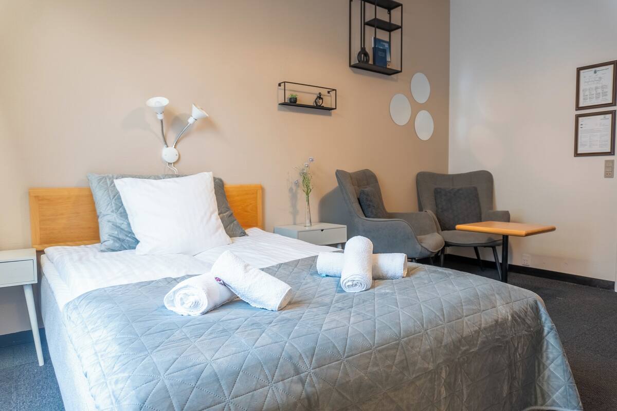 Una habitación de hotel con cama, mesita de noche, una silla, una pequeña mesa y un estante en la pared.