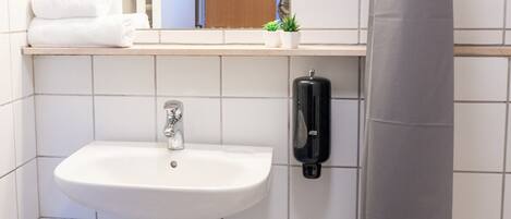 A bathroom with a white sink, a mirror, a towel rack with folded towels, a soap dispenser, and a small potted plant.