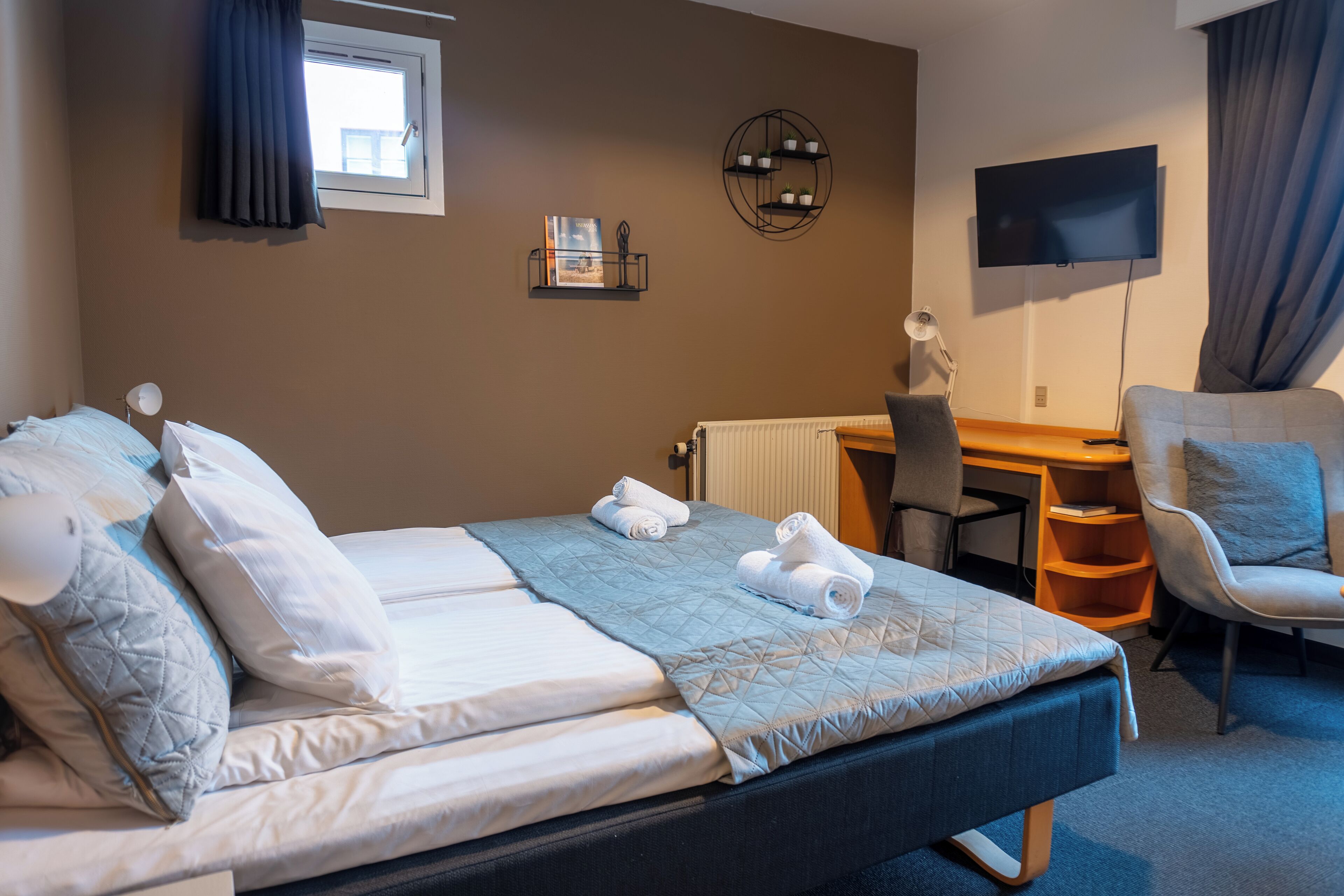 A hotel room with a bed, a desk, a chair, a TV, and a shelf with decorative items.