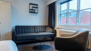 A modern living room with a grey sofa, a round coffee table, and a black armchair. There is a window with a view of a brick building outside. - Hotel Assens (Assens)