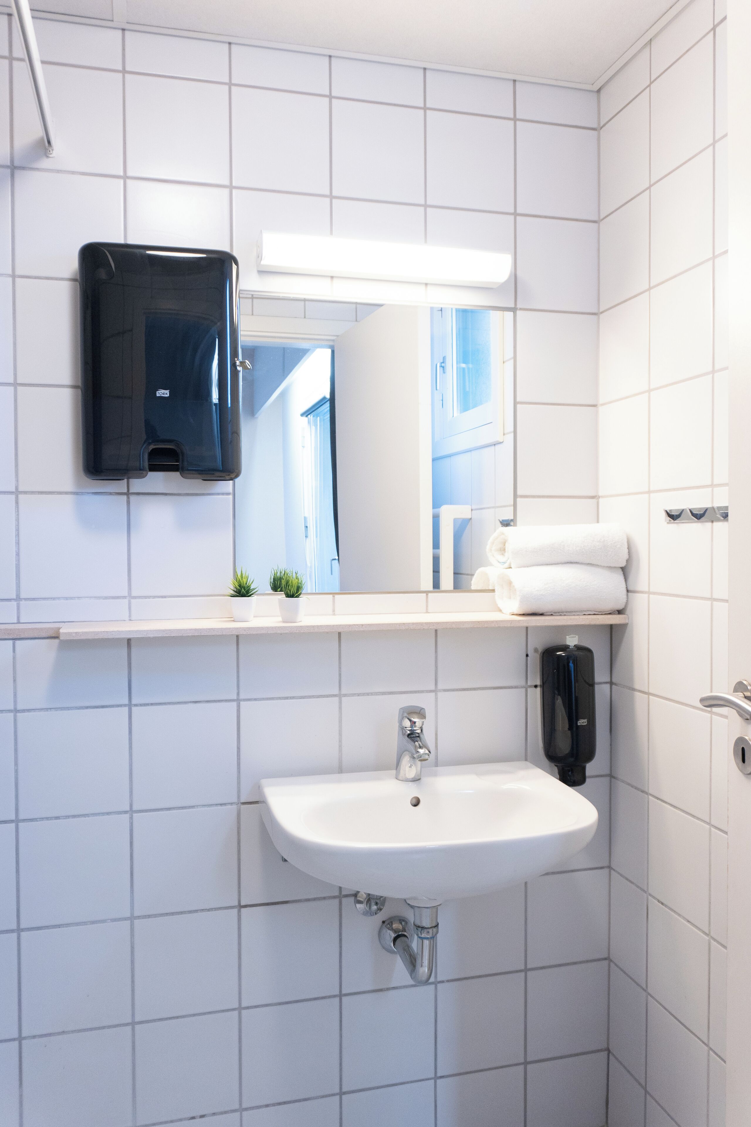 A bathroom with white tiles, a sink, a mirror, a towel rack, and a soap dispenser.