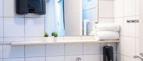 Un baño con azulejos blancos, un lavamanos, un espejo, un perchero para toallas y un dispensador de jabón.