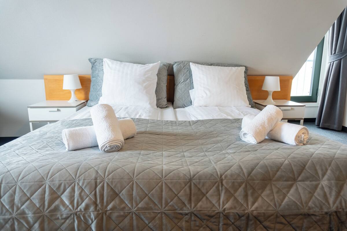 Una cama bien tendida con un cabecero acolchado, dos almohadas blancas y dos toallas enrolladas.
