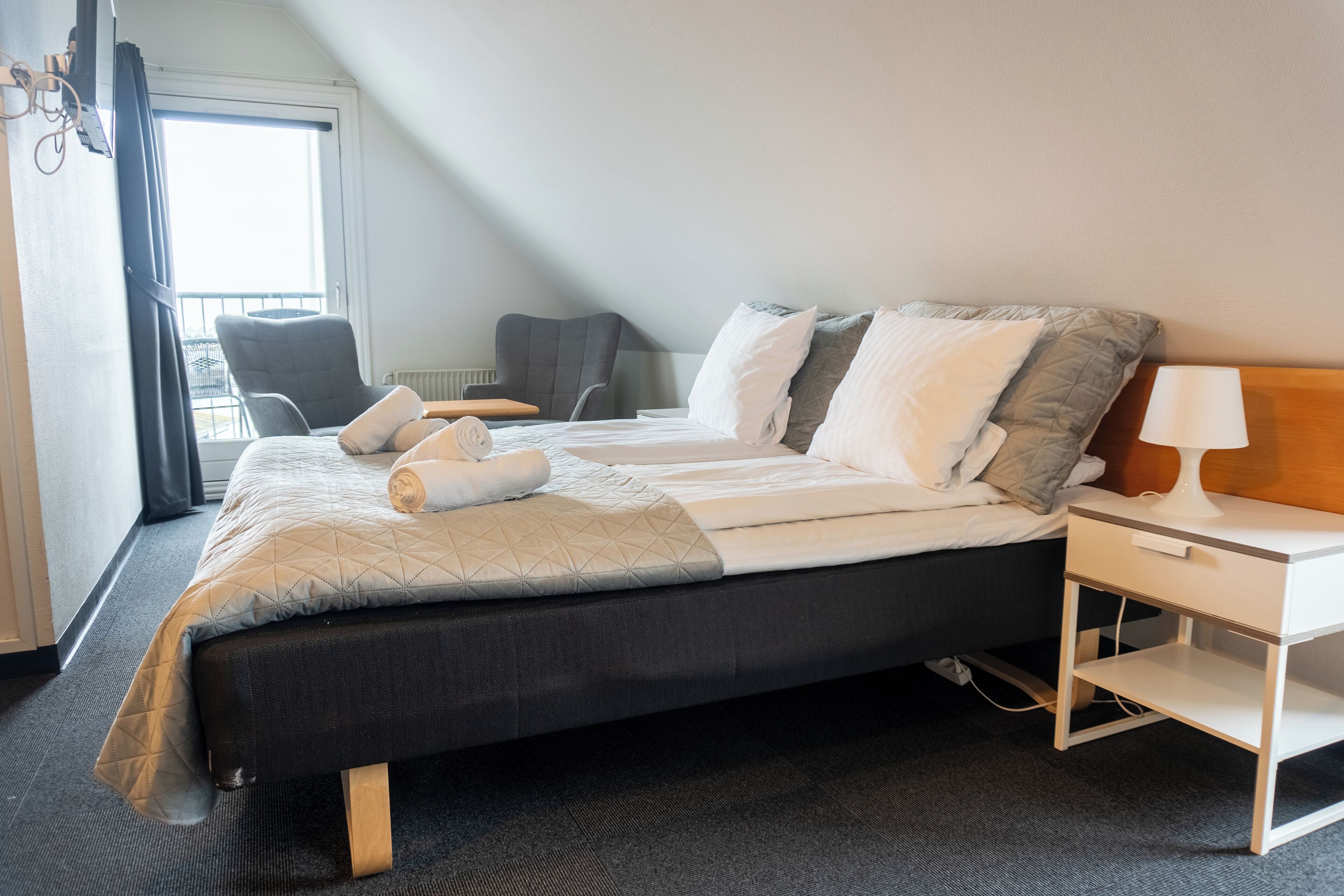 Habitación de hotel moderna con una cama grande, dos mesitas de noche, una silla y vistas al exterior por una ventana.