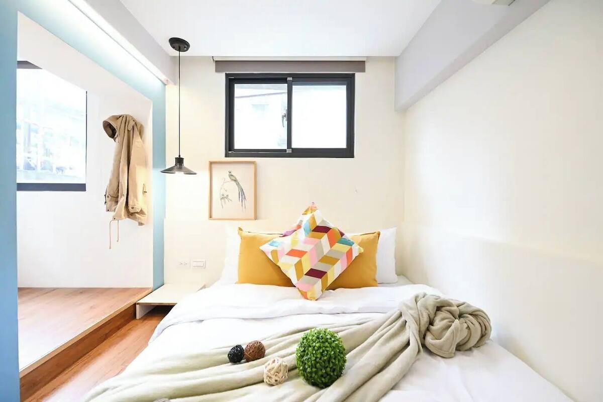 A bedroom with a bed, a window, a hanging coat, and a framed picture on the wall.